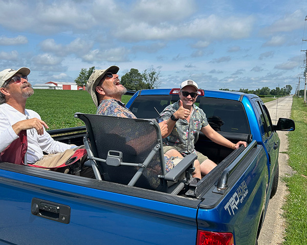 Three people sitting in a blue truck bed, two smiling and one giving a thumbs up.