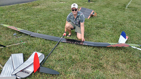 Man kneeling by large black model glider on grass, wearing a cap and sunglasses.