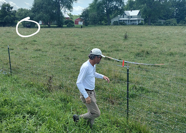 Man wearing a cap walking by a fence in a grassy field with trees and a house in the background.