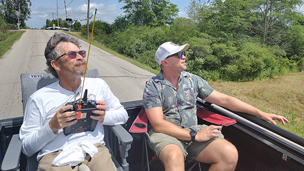 Two men sit in a vehicle, one holding a remote control, looking towards the sky.