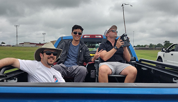 Three men in a truck bed, one holding a device, on a cloudy day.