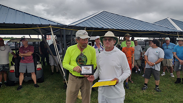 Man in hat receives an award from another man outdoors under a pavilion.