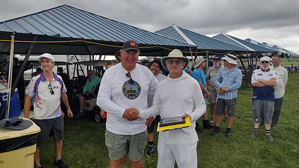 Two men in hats shaking hands, surrounded by a group at an outdoor event.