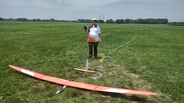 Person with hat stands on grassy field holding a remote near an orange glider.