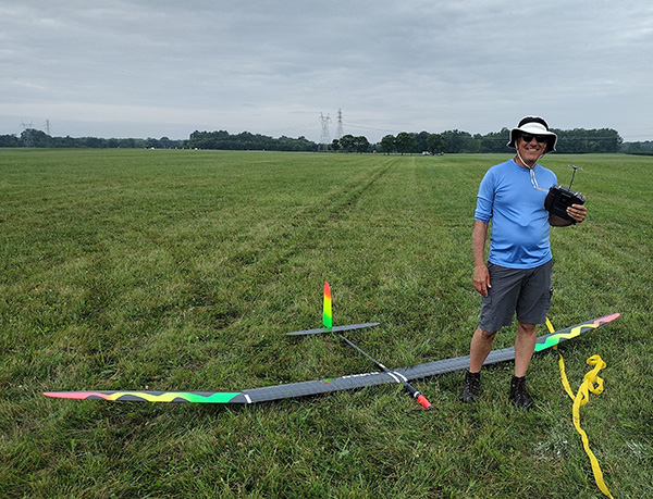 Man with hat stands next to a large model glider on a grassy field.