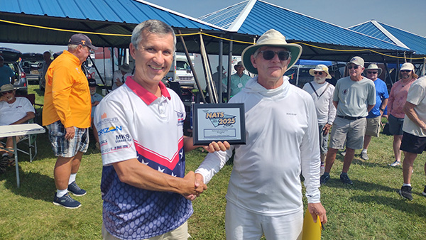 Two men shake hands, one holding a framed certificate, in an outdoor gathering.