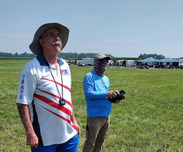 Two men in a grassy field, one holding a remote control, blue sky above.