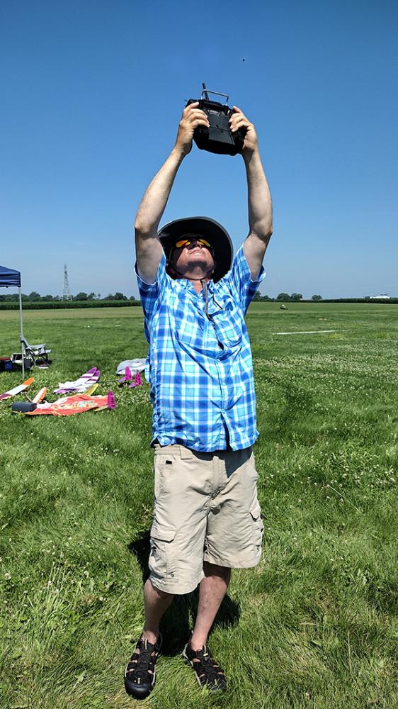 Man in blue plaid shirt and hat flies drone in sunny field.