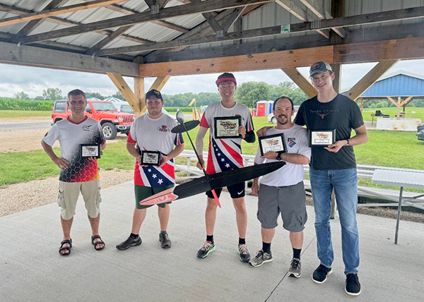 Five men holding plaques under a pavilion, smiling, with a countryside background.