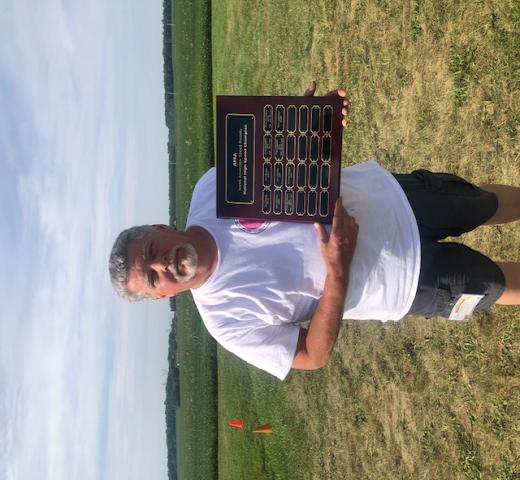 Man in white shirt holding a plaque, standing on grass with fields in the background.