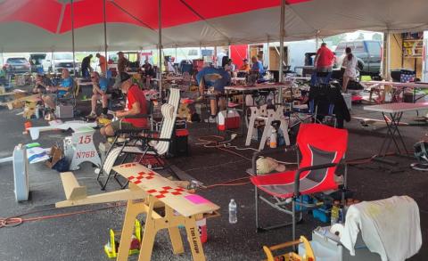 Outdoor event under a large tent with tables, chairs, and equipment scattered around.