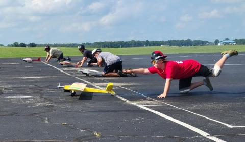Model planes being launched on a paved runway by people kneeling, under a blue sky.