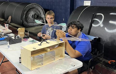 Steven Griggs and Dev Goda prepare their models for competition. Photo credit: Nick Ray Two people assemble a model aircraft at a table, large rolls in the background.