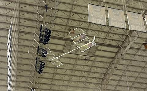 Model airplane flying indoors near a high ceiling with lights and signs.