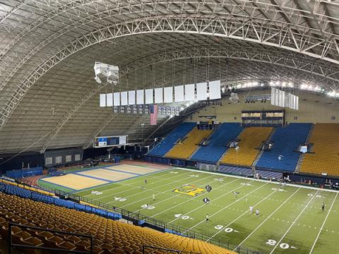 Indoor football stadium with players on the field and rows of blue and yellow seats.