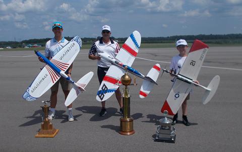 Three people outdoors with large model airplanes and trophies.