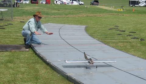 Man guiding small model airplane on a runway in a grassy field.