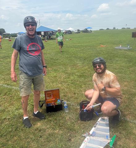 Two people with helmets outdoors, assembling a model airplane on grassy field.