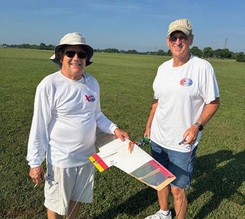 Two men smiling, holding a model glider on a grassy field.