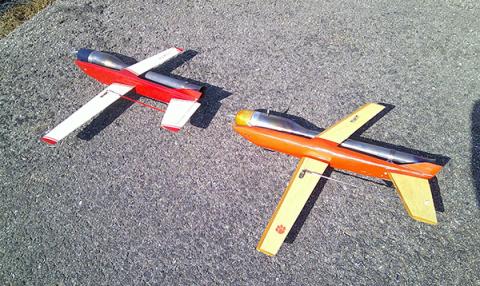 Two model airplanes on pavement, one red and white, the other orange and yellow.