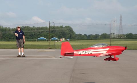 Red model plane flying low near a person on an airfield.