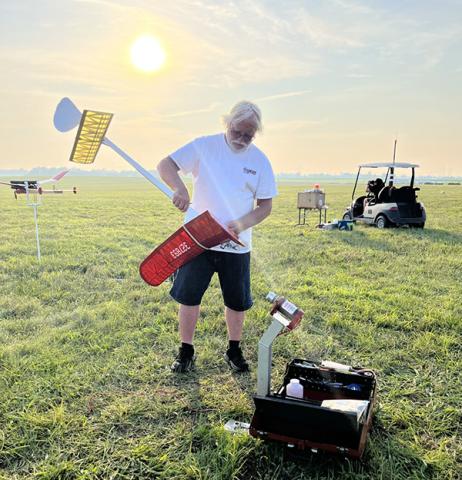 Man assembling red and white model plane on grassy field at sunrise.