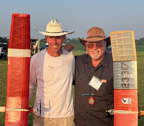 Two men smiling, wearing hats, stand outdoors holding model planes.