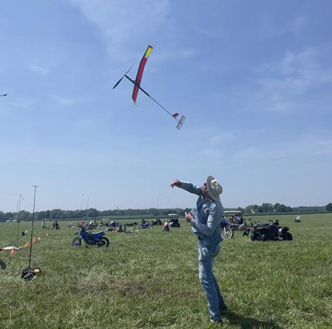 Man launching model glider in grassy field under clear blue sky.