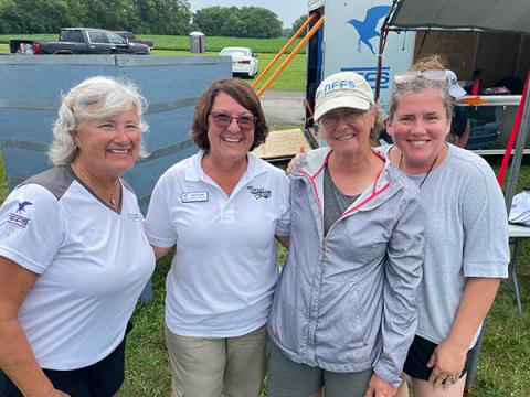 Four women smiling outdoors, wearing casual summer clothing.
