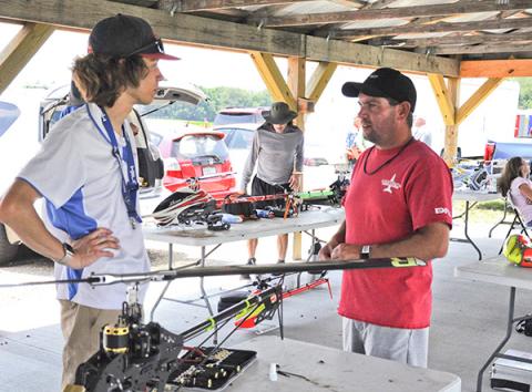 Two men discuss under a pavilion beside RC helicopters on tables.