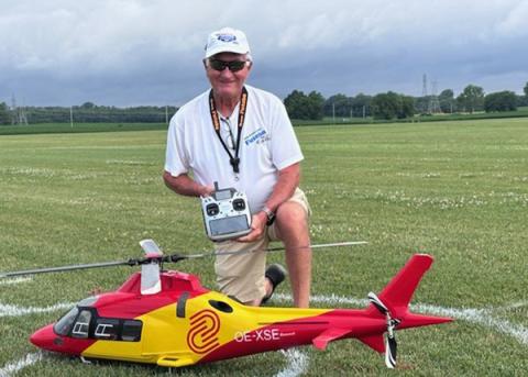 Man kneels on a field, holding a remote control next to a red and yellow model helicopter.