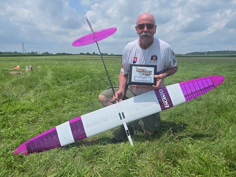 Man kneeling on grass with a model glider and holding a plaque.