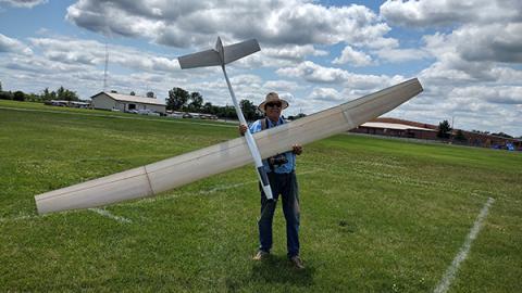 Man holding large model airplane on grassy field, cloudy sky overhead.