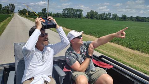 Two people in a truck bed, one holding a device, both looking and pointing at the sky.