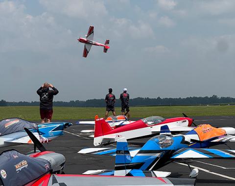 Aerobatic planes on a tarmac, one in flight, with spectators watching.