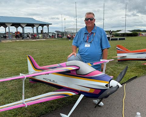 Man posing with colorful model airplane on grassy field.