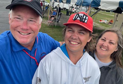 Three people smiling under blue and white tents at an outdoor event.
