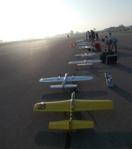 Row of model airplanes on a runway at sunrise, with people prepping nearby.
