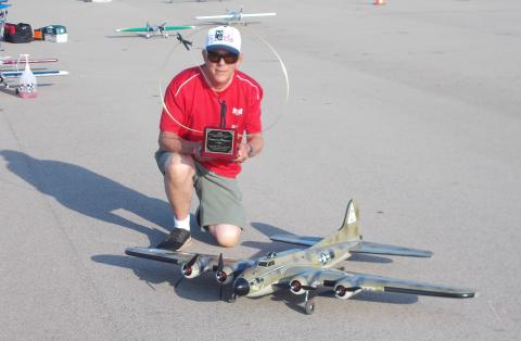 Man kneeling with award, posing next to a model airplane on a sunny airfield.