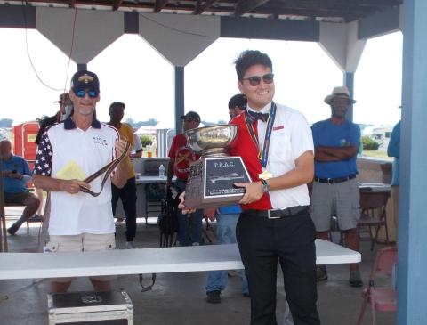 Young man holding a trophy, smiling; other people present in a pavilion setting.