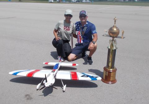 Two people kneel by a large trophy and model airplane on a tarmac.