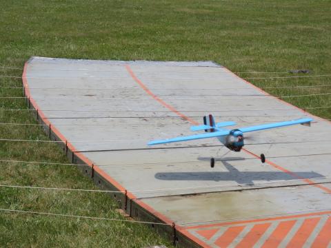 A small blue model plane taking off from a wooden runway on grass.
