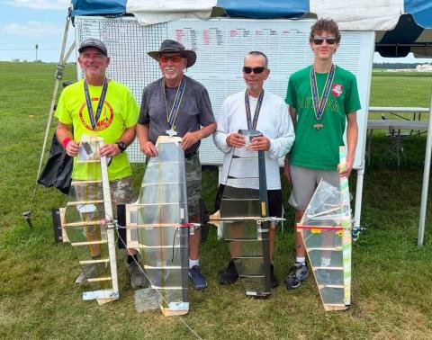 Four men standing on grass, each holding a model airplane, smiling for a photo.