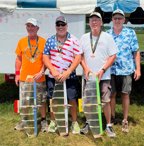 Four men standing with model gliders, wearing medals and casual clothing, outdoors under a tent.