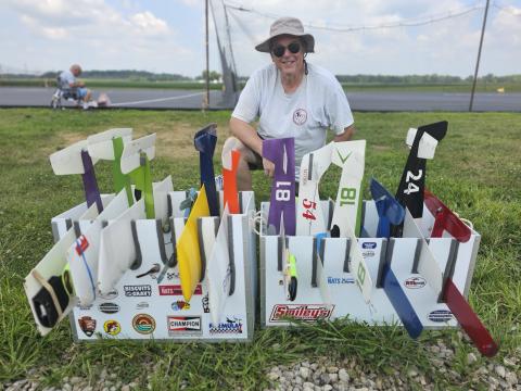 Man in sunhat smiling beside model planes on grass.