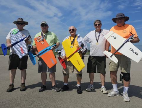 Five men with remote control planes, smiling, standing on pavement under clear sky.