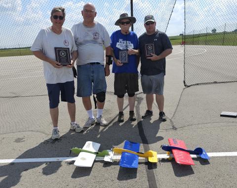 Four men holding plaques stand beside colorful model airplanes on pavement.