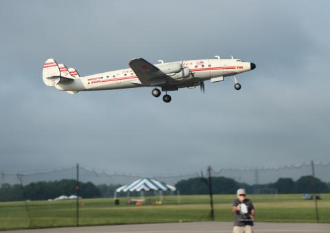 RC plane flying low over a field, man controlling it on the ground.