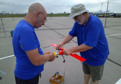 Two men in blue shirts fixing a red model airplane outdoors.