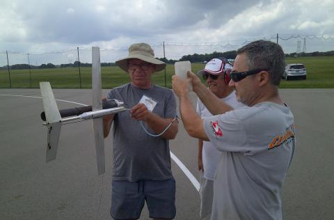 Three men on an airfield inspect a model rocket, holding fuel container and tubing.